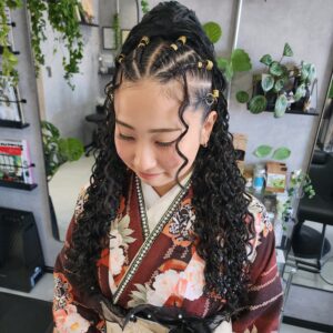 ボリュームのあるスパイラルカールをベースに、フロントとトップに多数の小さなゴールドのリングやチューブ状のヘアアクセサリーが固定された複数のコーンロウ（編み込み）を施したアップスタイル。顔の周りには、細いスパイラルカールの毛束が後れ毛として垂れ下がっている