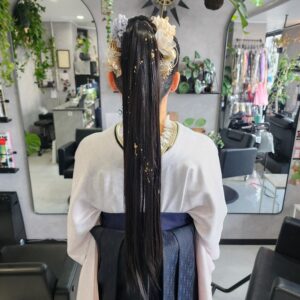 和装（白い上衣と紺色の袴）姿の人物を後ろから捉えた写真。腰まである非常に長い黒髪が、高めの位置でポニーテールにまとめられている。髪の表面には繊細な金箔が散りばめられ、ポニーテールの根元は、くすんだ青、ベージュ、ゴールド系のドライフラワーと、ゴールドの髪飾りで華やかに飾られている。背景は、鏡と多数の観葉植物が配置されたスタイリッシュな美容室の内装。卒業式や成人式を連想させる、現代的で華やかな和装ヘアスタイル。