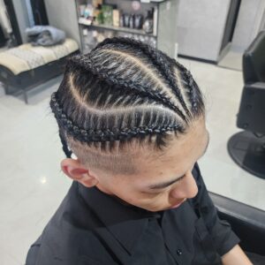 男性の複雑なコーンロウ（編み込み）ヘアスタイルのクローズアップ写真。斜め上からの視点で、頭頂部には前髪のヘアラインから後方へ向かう複数の精巧な編み込みが、前方は密に、後方はより広いセクションでV字型やジグザグの複雑なパターンを描きながら整えられている。側面と後頭部は短く刈り込まれたフェードカットで、境界がはっきりしている。