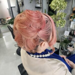 鮮やかなピンク色のショートボブを、ふんわりとしたカールアップスタイルにアレンジした女性の後ろ姿。豊かなカールを重ねて後頭部でねじりながらまとめ上げ、襟足はスッキリと、サイドはカールした後れ毛を残しています。