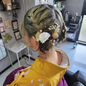 鮮やかな黄色の和装（振袖または訪問着）に合わせた、複雑で華やかなヘアセットの後ろ姿。彼女の髪は、ダークグリーンからオリーブグリーンへのグラデーションカラーで、ルーズな編み込みを組み合わせて低い位置で2つの小さなシニヨンにまとめられています。髪全体に繊細な金箔（ゴールドフレーク）が散りばめられ、後頭部には白い造花、パールのクラスター、大きなゴールドのラメボール、小さなゴールドのピンなどの豪華な髪飾りが組み合わさっています。
