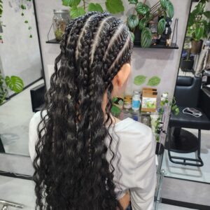 スタイリッシュな美容室の店内で撮影された、女性の後ろ姿。頭頂部と側面に沿って約8本の太いコーンロウが編み込まれており、後頭部の中央で終了して、残りの非常に長い髪は豊かなボリュームのあるディープカールとして下ろされている。彼女は白いTシャツを着用しており、背景には鏡、観葉植物（モンステラ、スイカペペロミア）、ヘアケア製品の棚が見える。コーンロウのタイトさとカールのルーズさが対照的なハーフアップスタイル。
